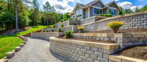 A beautifully designed tiered retaining wall with lush greenery and a modern home in the background, showcasing expert landscaping and hardscaping.